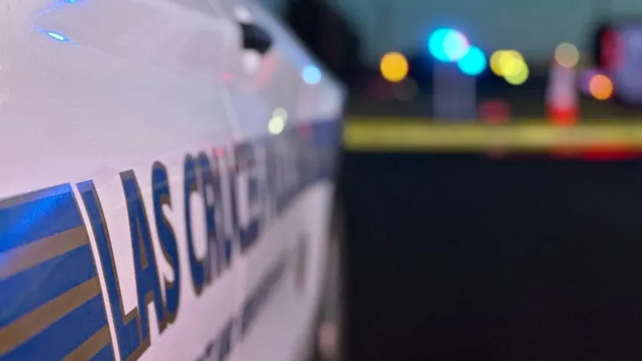 Close-up of a Las Cruces Police Department vehicle with blurred flashing lights and yellow crime scene tape in the background.