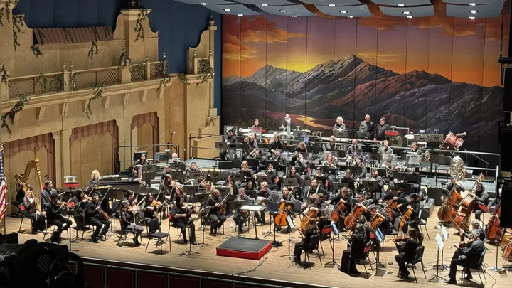 The El Paso Symphony Orchestra performs onstage at the Plaza Theatre, with musicians arranged in sections beneath a backdrop depicting mountains at sunset.