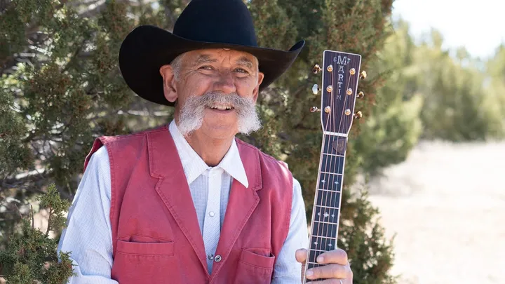 Singer-songwriter and rancher Randy Huston stands outdoors wearing a black cowboy hat and red vest, smiling while holding his Martin guitar near juniper trees in rural New Mexico.