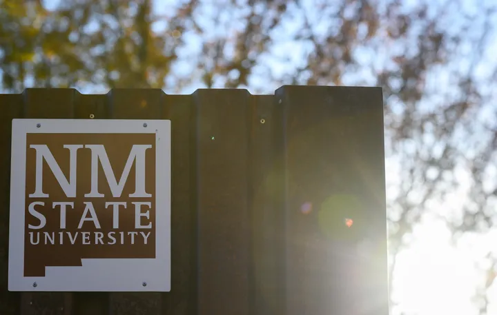 A metal sign displays the NM State University logo in white against a brown background, illuminated by morning sunlight with blurred trees in the distance.