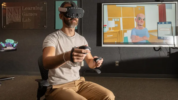 A man wearing a virtual reality headset and hand controllers participates in a nutrition-training simulation at NMSU’s Learning Games Lab, with an animated patient shown on a nearby screen.