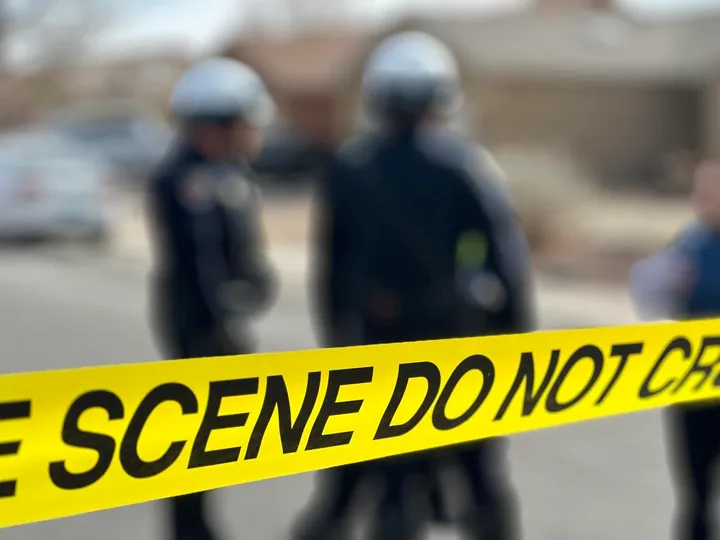 Yellow police tape reading “Crime Scene Do Not Cross” in focus, with blurred law enforcement officers standing in the background.