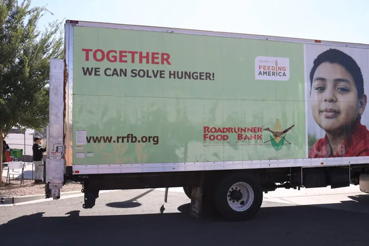 A Roadrunner Food Bank truck with the message “Together we can solve hunger!” and a photo of a child is parked at a community food distribution site during a Feeding America partner event.