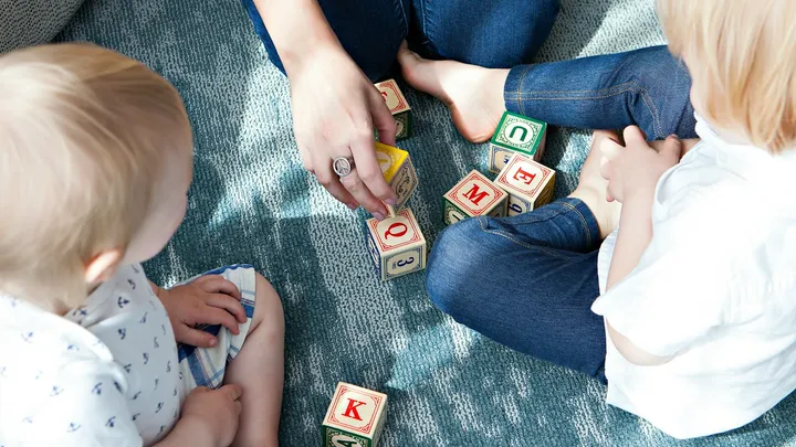 Two small children sit on a rug playing with colorful alphabet blocks as an adult arranges the pieces beside them.