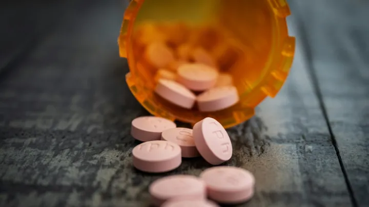An open orange prescription pill bottle tipped over on a wooden surface with pink tablets scattered in front of it.