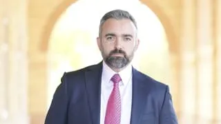 Attorney General Raúl Torrez, in a navy suit, white shirt and pink tie stands facing the camera inside a softly lit arched corridor with a bright background.