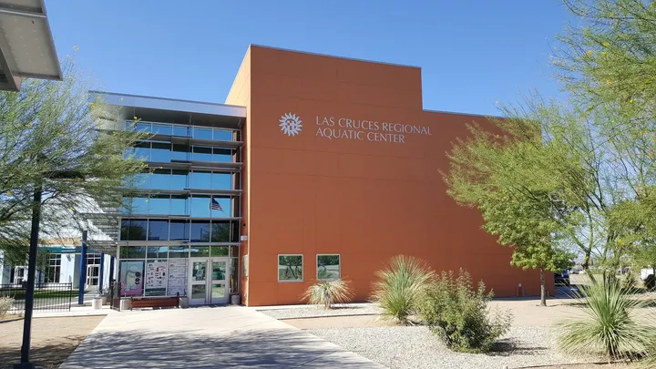 Exterior of the Las Cruces Regional Aquatic Center on East Hadley Avenue, showing its orange and glass facade under a clear blue sky with desert landscaping in front.