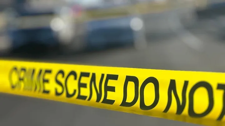 Close-up of yellow police crime scene tape stretched across a parking lot, with blurred vehicles visible in the background.