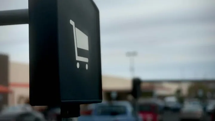 A shopping cart symbol on a sign in a parking lot outside a retail store, with cars and a cloudy sky in the background.
