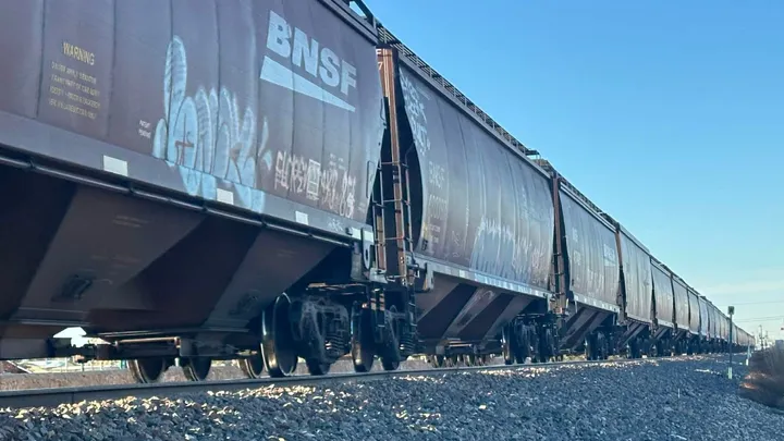 A line of BNSF freight train cars travels north along railroad tracks near south Main Street in Las Cruces under a clear blue sky, with gravel embankment and graffiti visible on the cars.