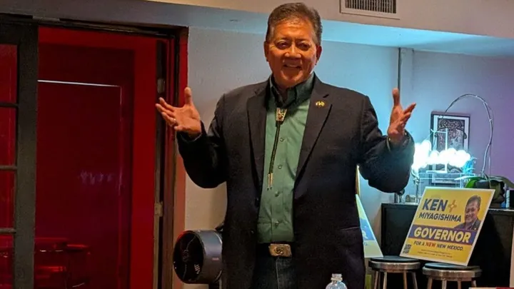 Former Las Cruces Mayor Ken Miyagishima speaks to supporters indoors, standing with his arms raised near campaign signs during his run for New Mexico governor.