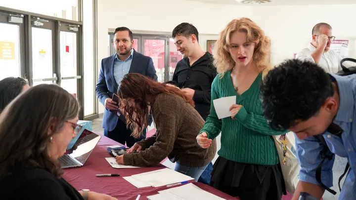 People check in at a registration table during an NMSU event, with students filling out forms and staff seated behind laptops in a busy lobby area.