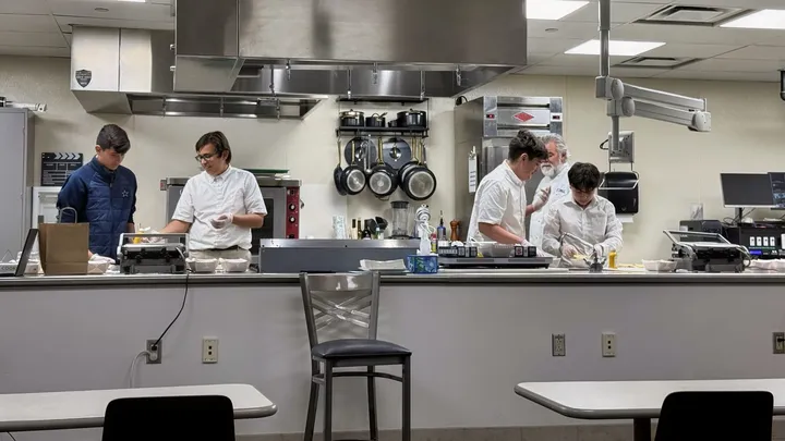 Middle school students work alongside instructors in DACC’s commercial kitchen, preparing food at stainless steel workstations with pots, pans and cooking equipment visible.