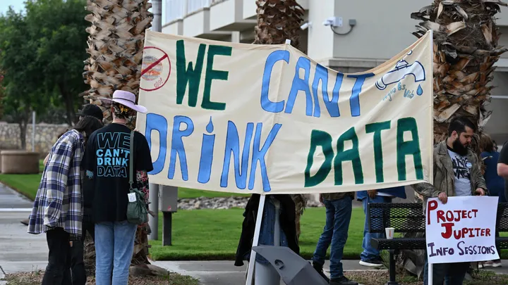 Protesters stand outside a building holding a banner reading “We can’t drink data,” with additional signs opposing Project Jupiter visible nearby.