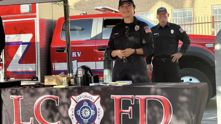 Two Las Cruces Fire Department firefighters stand behind an LCFD information table, with a fire engine parked behind them at an outdoor community event.