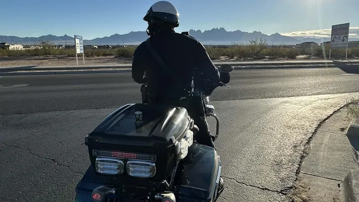 A Las Cruces police officer on a motorcycle watches traffic on Red Hawk Golf Road with the Organ Mountains visible in the distance.