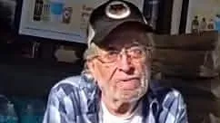An older man with gray hair and glasses wears a black baseball cap, blue plaid shirt and white T-shirt while sitting indoors with his arms crossed. He uses a cane to help walk.