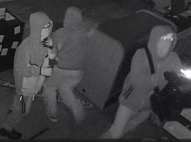 Grainy black-and-white surveillance image shows several masked individuals wearing hooded sweatshirts inside a building during a nighttime burglary.
