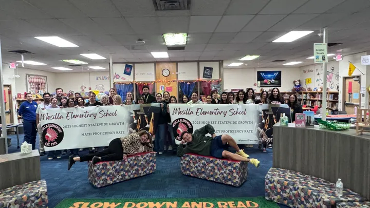 A large group of Mesilla Elementary staff pose in the school library with two banners celebrating statewide growth in literacy and math, with two staff members playfully posing in front.
