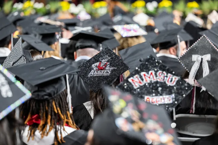 Graduates wearing black caps and gowns sit closely together during an NMSU commencement ceremony, with decorated mortarboards visible in the crowd.