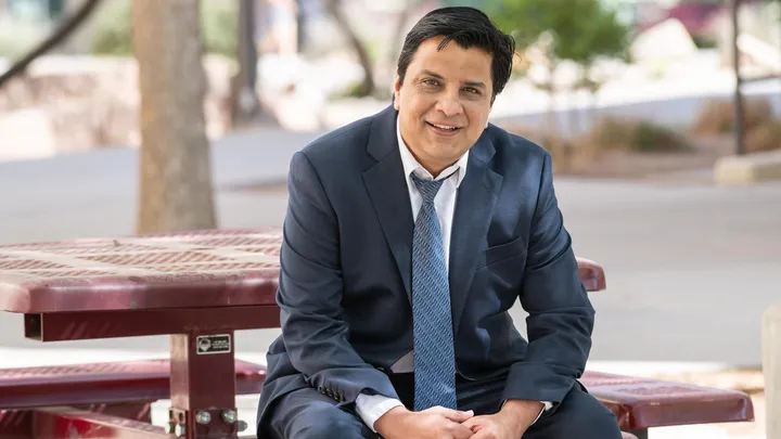 Jagdish Khubchandani, in a navy suit and tie, sits at an outdoor picnic table and smiles toward the camera on the New Mexico State University campus.