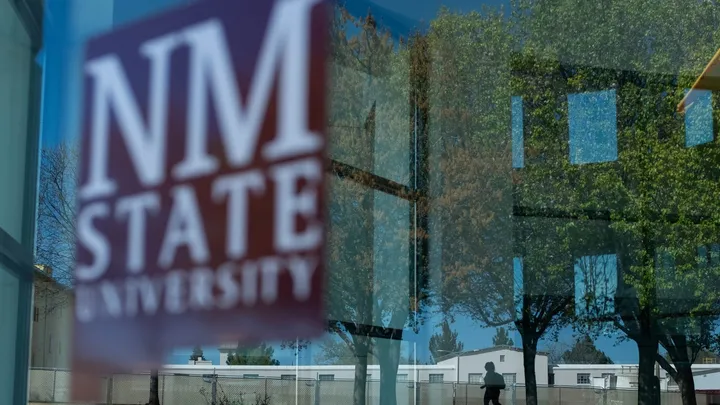 A window reflects trees and campus buildings near New Mexico State University, with the NMSU logo visible on the glass.