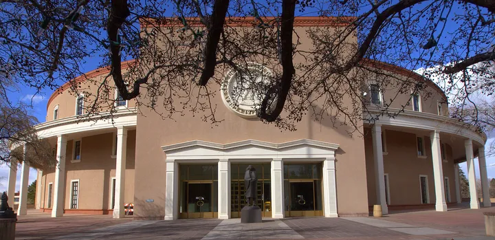The New Mexico State Capitol, known as the Roundhouse, is seen in Santa Fe with its circular exterior and columns framed by tree branches in the foreground.