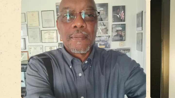 Volunteer ombudsman Kevin Thompson stands indoors wearing glasses and a button-down shirt, with framed certificates and military photos displayed on the wall behind him.