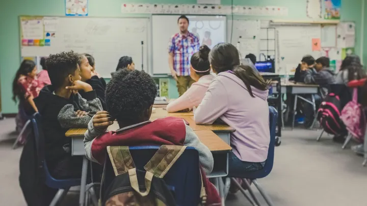 A classroom full of students sits at desks facing a teacher at the front of the room during a lesson.