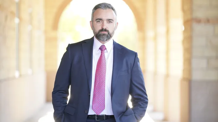 New Mexico Attorney General Raúl Torrez stands in a sunlit corridor wearing a dark suit and pink tie, facing the camera.
