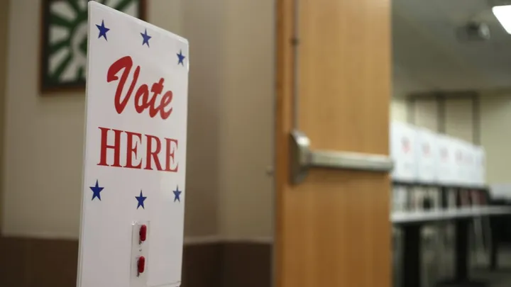 Vote Here sign stands inside a polling place, with voting booths visible in the background.