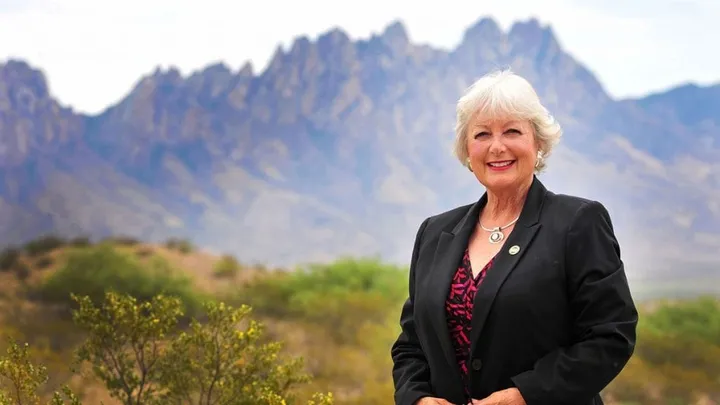State Rep. Joanne Ferrary stands outdoors in front of the Organ Mountains, wearing a black blazer and patterned top, with desert vegetation visible in the foreground.