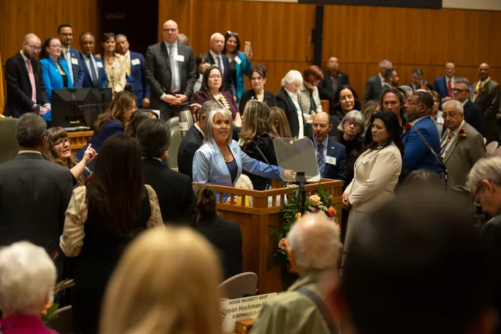 Gov. Michelle Lujan Grisham speaks at the podium in the New Mexico House chamber during the State of the State as lawmakers and guests stand around her.