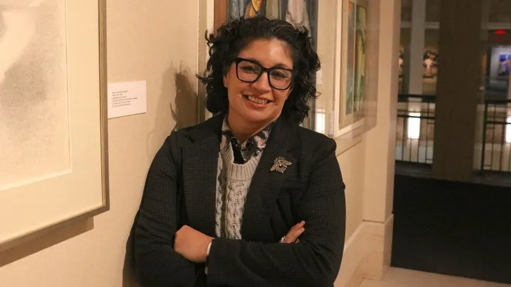 State Rep. Sarah Silva stands indoors in a gallery space with framed artwork on the walls behind her, smiling with her arms crossed.