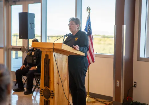 Sheriff Kim Stewart speaks at a podium in Sunland Park, with an American flag behind her and attendees seated nearby.