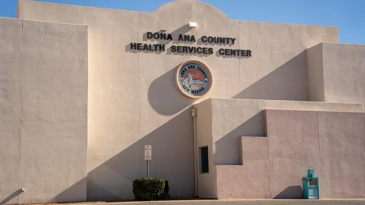 Exterior of the Doña Ana County Health Services Center building in Las Cruces, with the county seal mounted on the stucco facade beneath a clear blue sky.