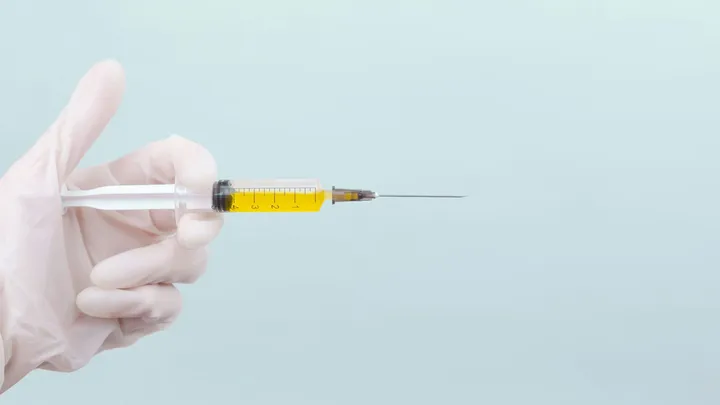 A gloved hand holds a syringe filled with yellow liquid against a light blue background.