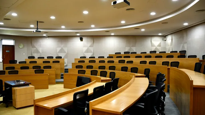 Empty courtroom with tiered seating, wood-paneled desks and a judge’s bench at the front.