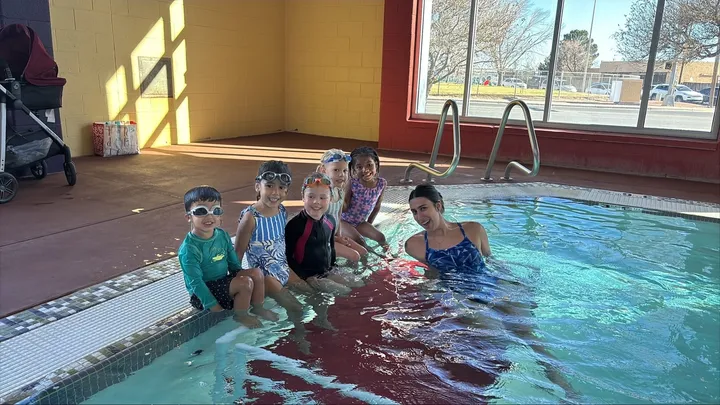 Children wearing swim goggles sit at the edge of an indoor pool with an instructor during a swim lesson at a Las Cruces aquatic facility.