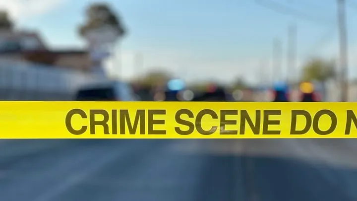 Yellow crime scene tape reading “Crime Scene Do Not Cross” stretched across a roadway with police vehicles and flashing lights blurred in the background.