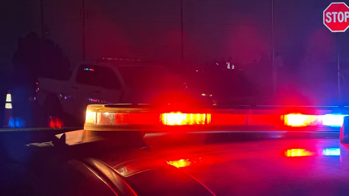 Red and blue emergency lights flash on top of a police vehicle at night while other law enforcement vehicles and traffic cones are visible in the background.