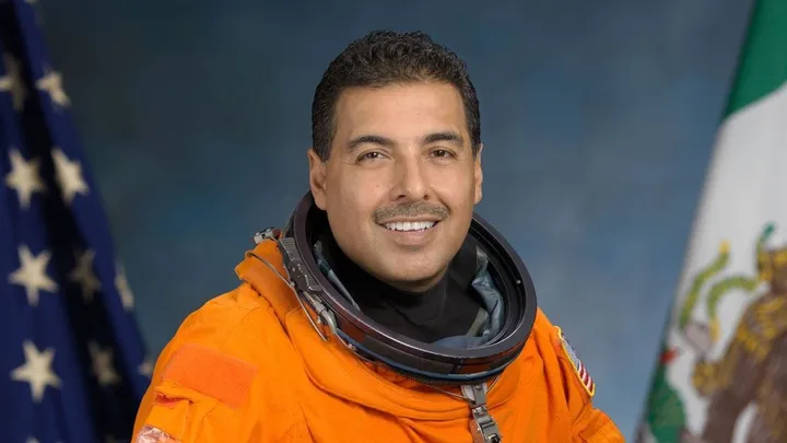 Portrait of José Hernández in an orange NASA astronaut suit with U.S. and Mexican flags in the background