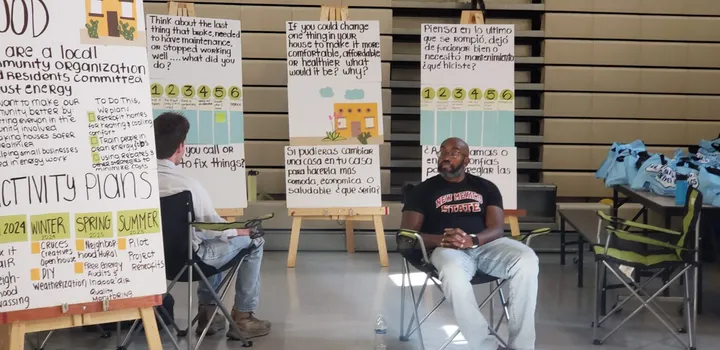 Residents sit in folding chairs during a community meeting in Las Cruces, with posters outlining housing, maintenance and energy improvement ideas displayed in English and Spanish