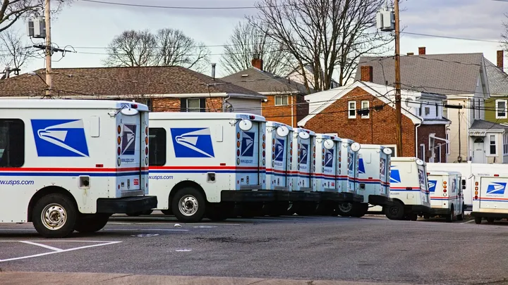 U.S. Postal Service delivery trucks are lined up in a parking lot near a residential neighborhood