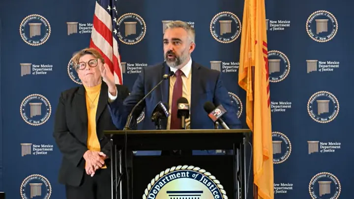 New Mexico Attorney General Raúl Torrez speaks at a podium during a press conference, with a woman standing behind him against a New Mexico Department of Justice backdrop.